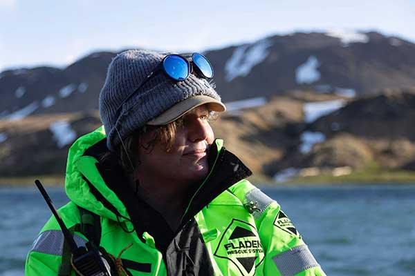 Katie Marx in a bright green jacket holds a black walkie-talkie, with a body of water and mountains in the background.