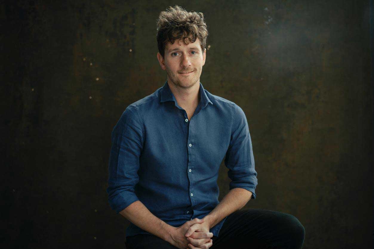 Robbie Arnott seated with hands clasped, wearing a blue shirt, against a dark textured background.