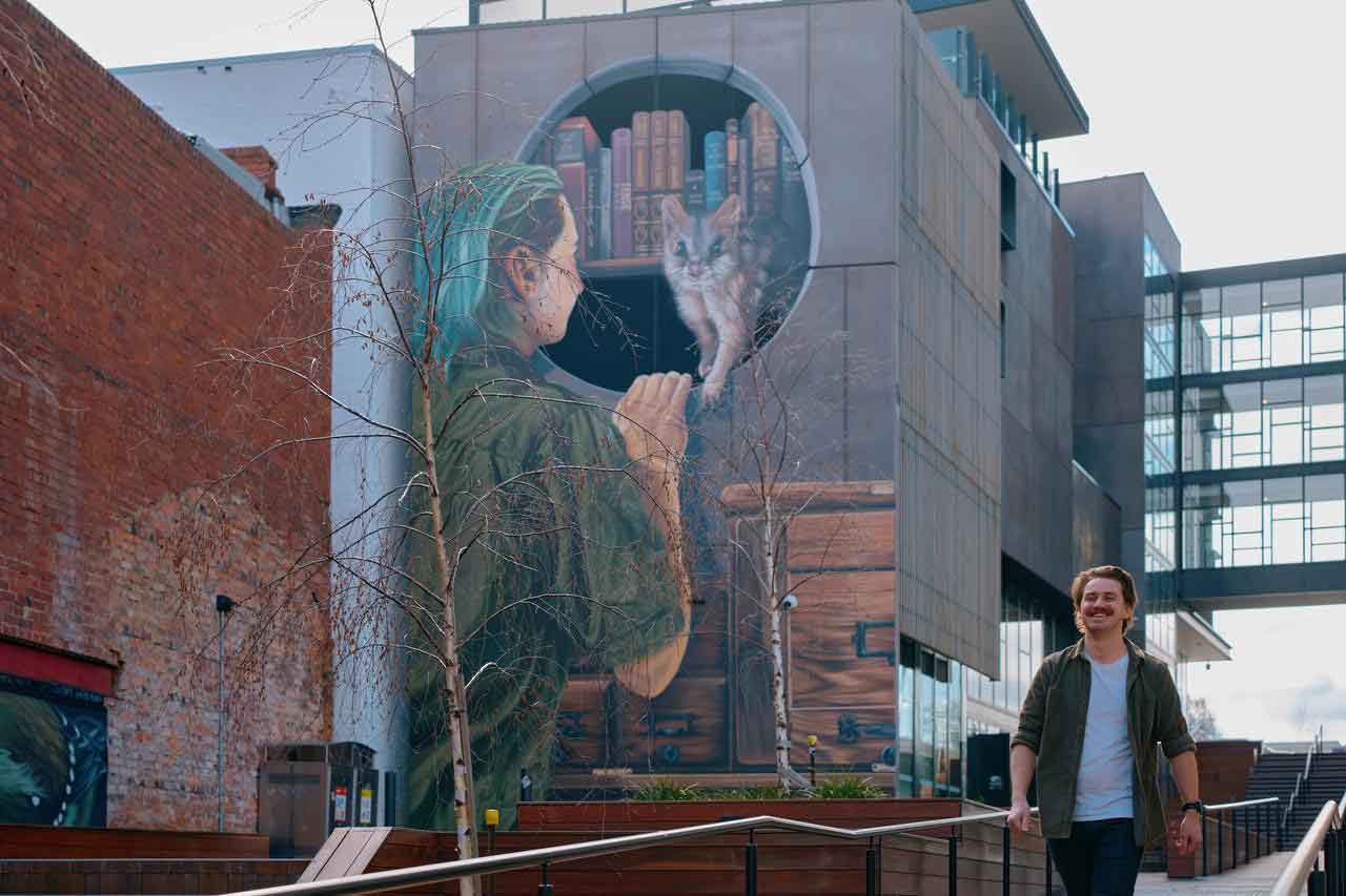 A mural on a building depicting a giant hand holding a pencil, with a cat peeking out from a hole in the wall, surrounded by books.