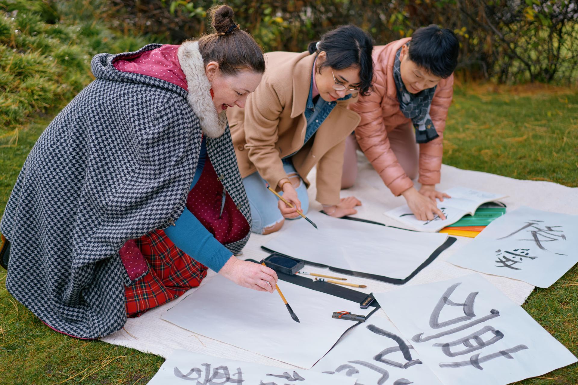 A female teacher kneeling on the grass in a garden setting with a male and female student painting Japanese characters