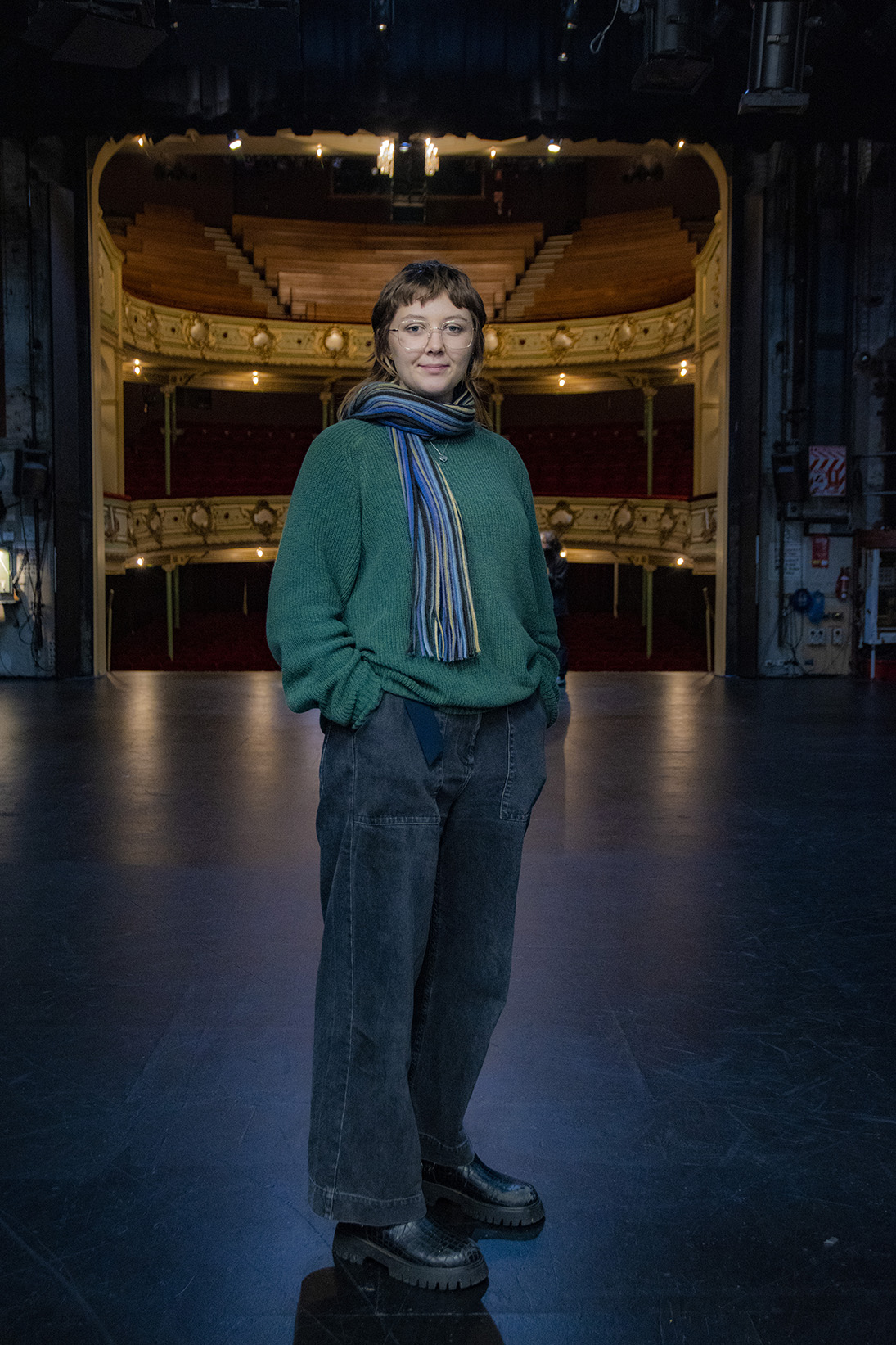 Young woman wearing a green jumper and blue jeans standing on the stage with the theatre behind her