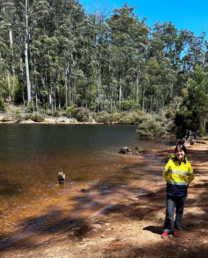 A tea tree stained river with the tall tress and a woman in an Entura yellow jumper standing on the bank