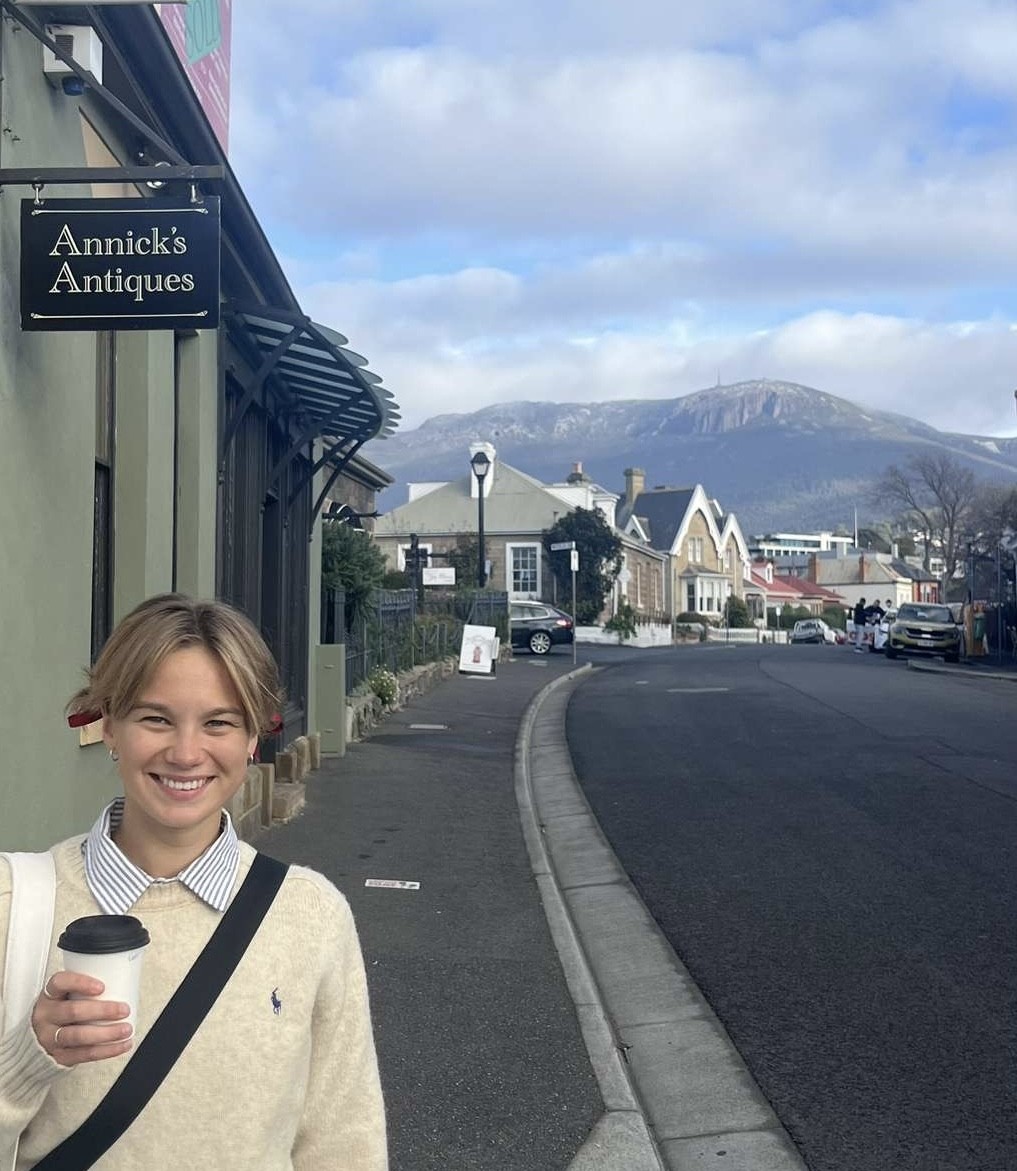 Imogen McMullen holds a coffee, standing in a Battery Point street, with kunanyi/Mt Wellington in the distance.