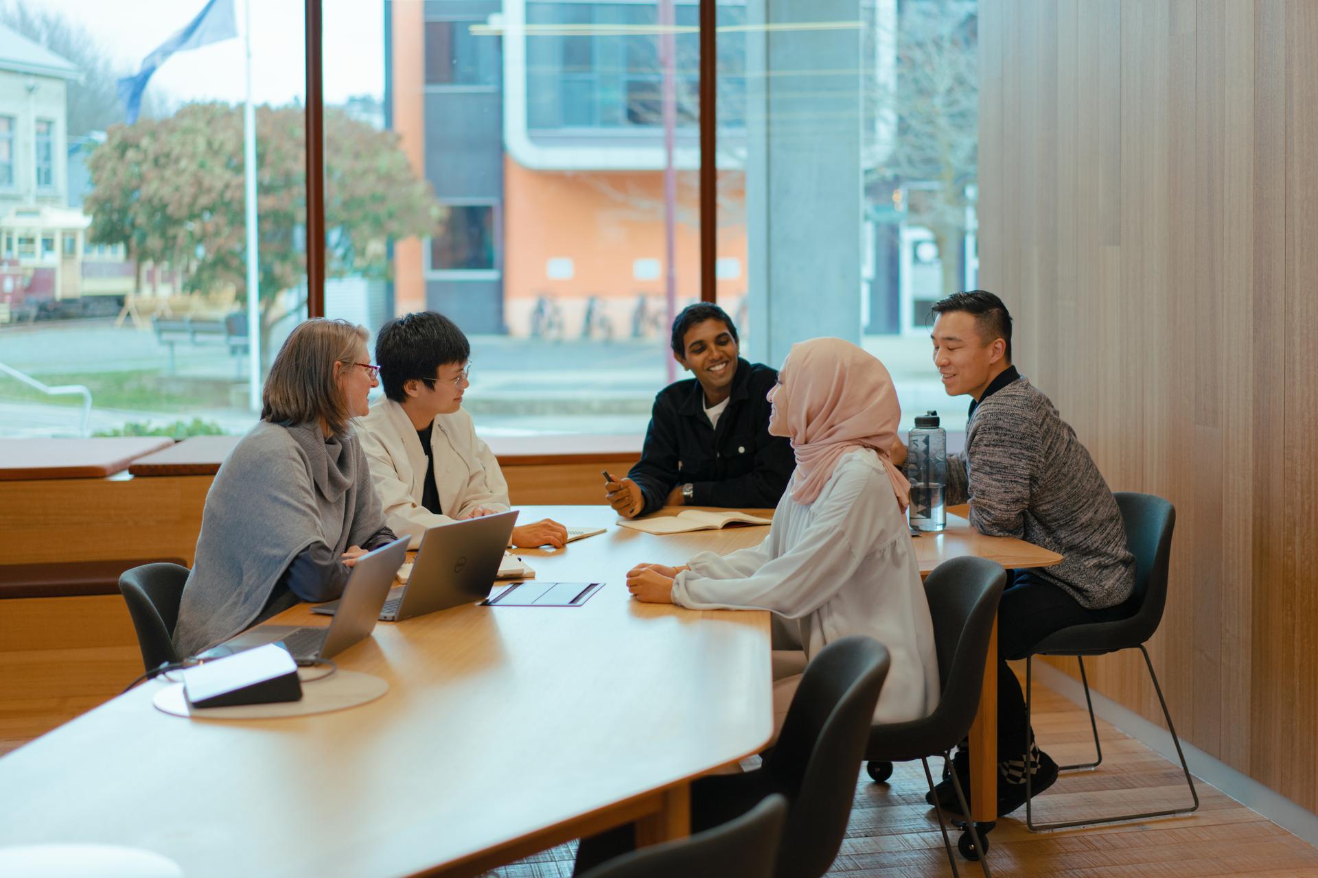 Two women and three men seating around a table neat to a window in a library 