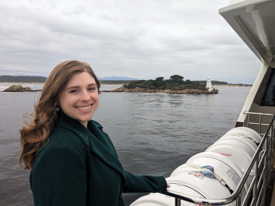 A young woman in a dark green coat on a boat with a lighthouse on an island in the background