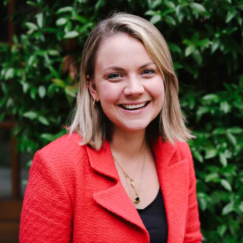 Cassandra Hugillwearing a red blazer stands in front of green foliage.