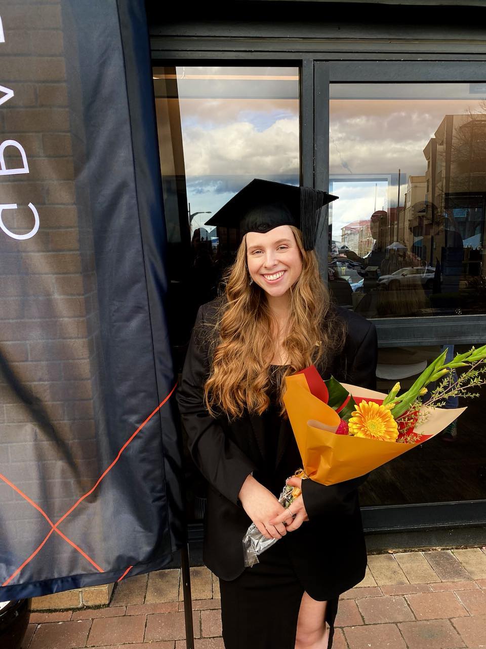 A young woman in a black graduating gown and mortar board with a large bouquet of flowers