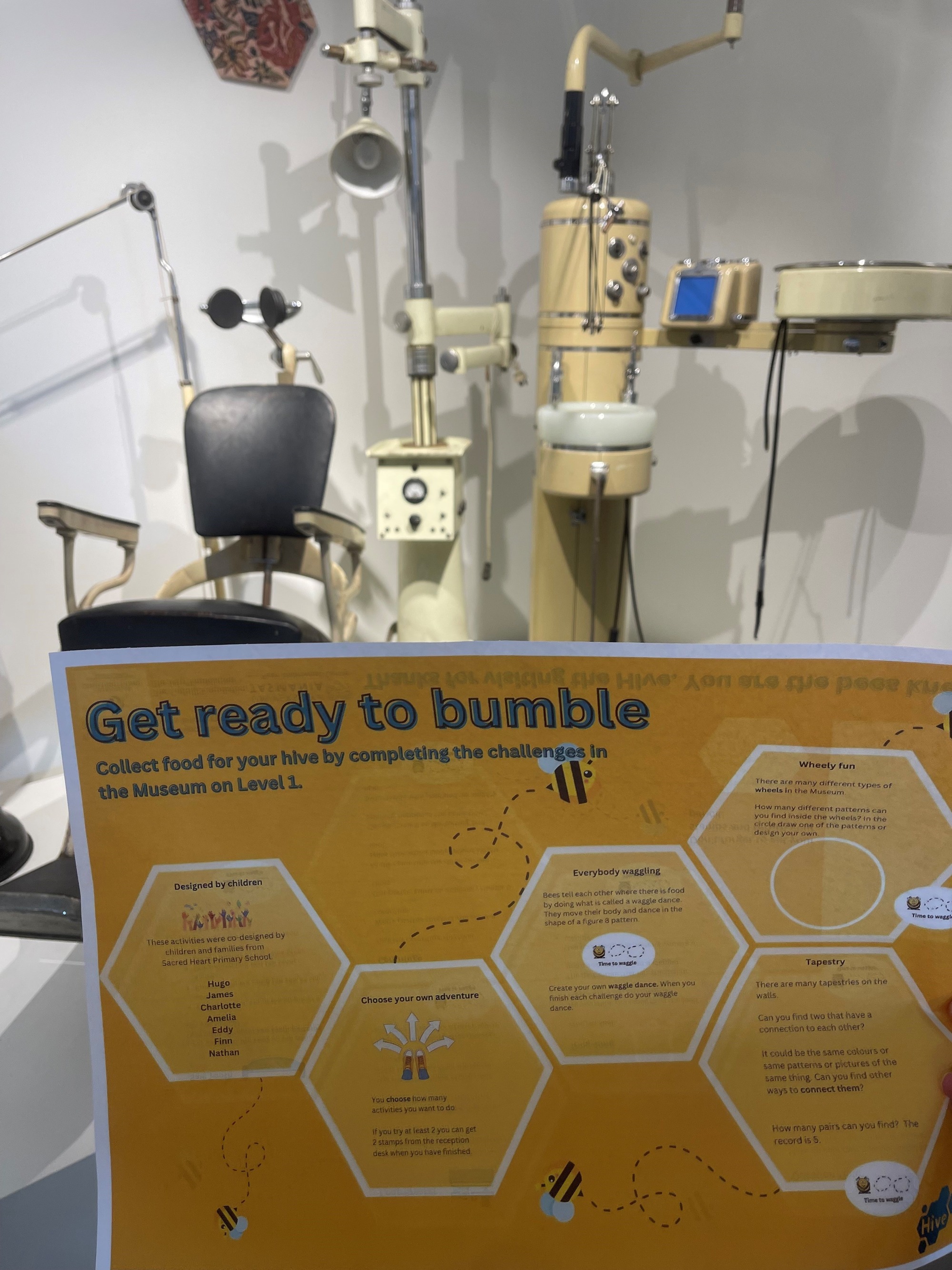 A close-up of a worksheet being held in someone's hand in front of a Museum exhibit. The worksheet is titled 'Get ready to bumble' and has honeycomb style hexagons laid out over a yellow background with pictures of bees flying from some of the shapes. Inside each hexagon is a challenge for children to undertake in the museum.