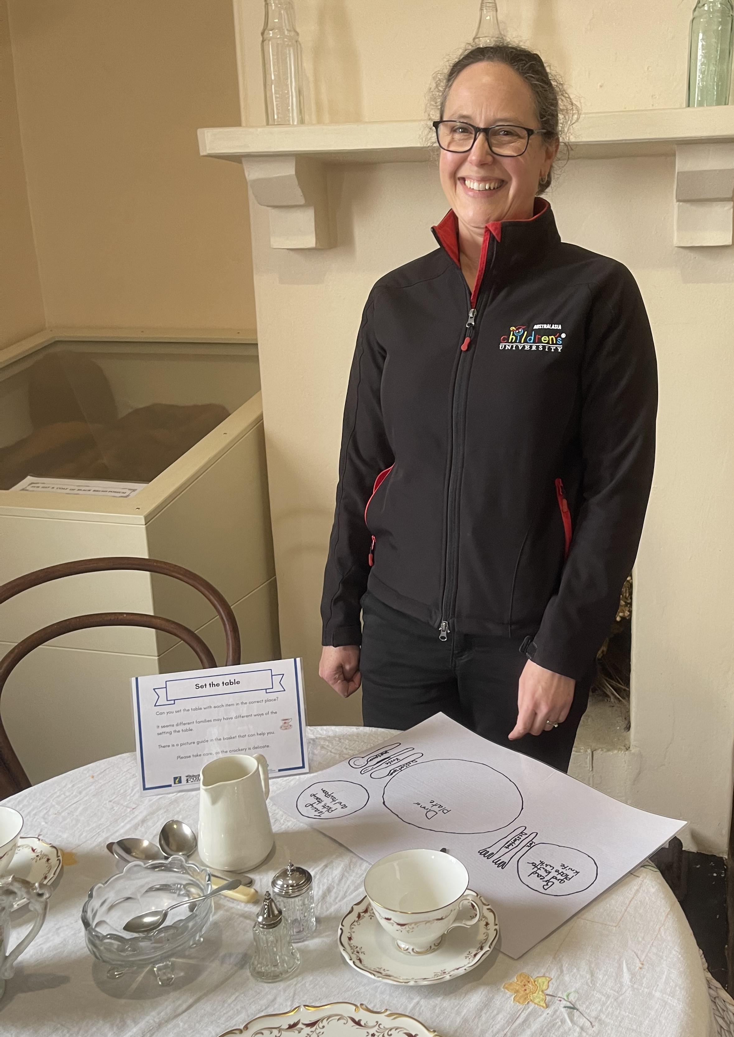 Claire Robertson stands in front of the 'set the table' activity at the Great Western Ties Visitor Centre. The table is covered in vintage plates and tea cups and saucers, and Claire is smiling and wearing a Children's University branded jacket.