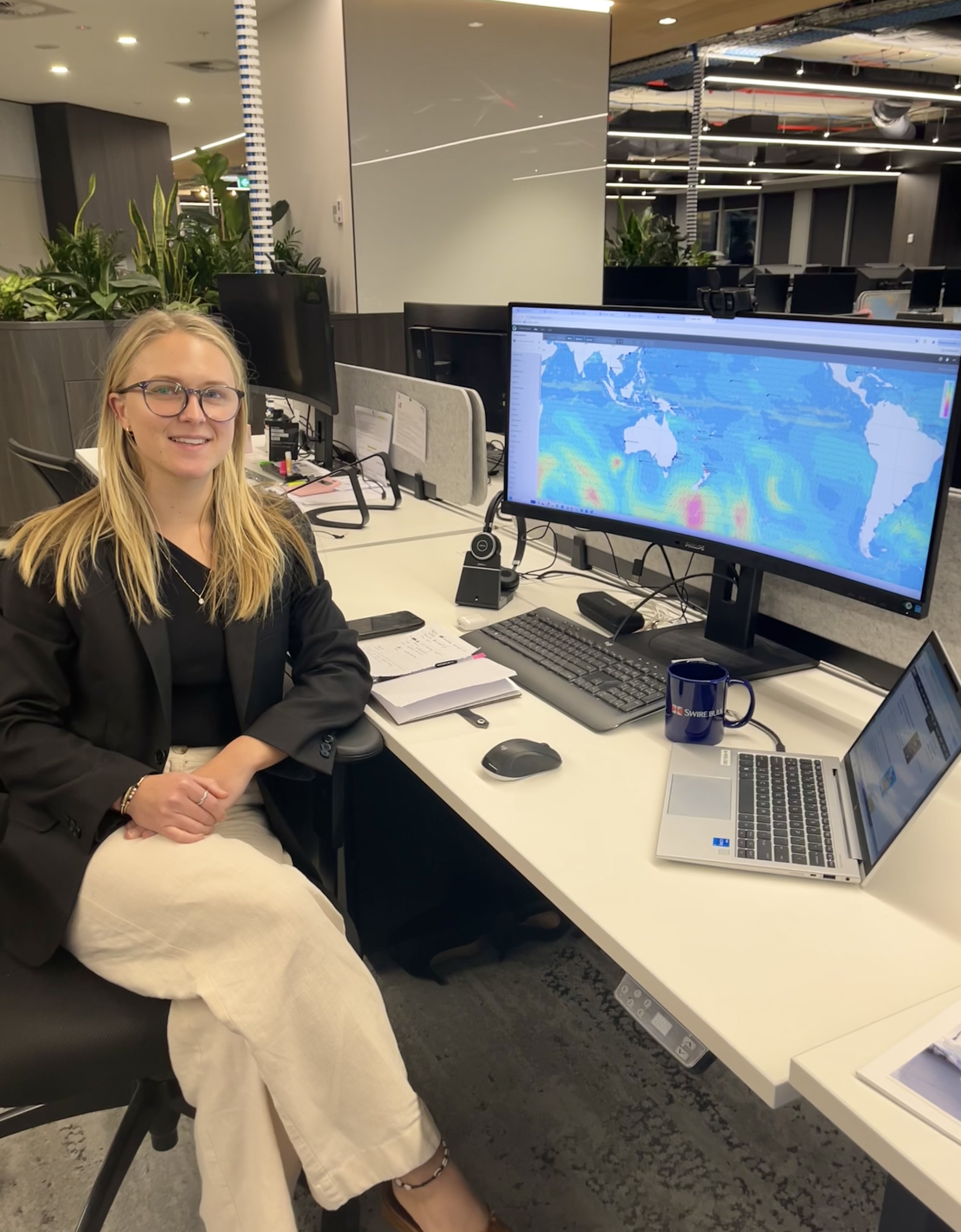 Australian Maritime College graduate Gabrielle Hall is seated at a desk in an open-plan office. She has shoulder-length blonde hair, glasses, and is wearing a black top and blazer, smiling at the camera. A computer sits on the desk, a colourful world map on the monitor.