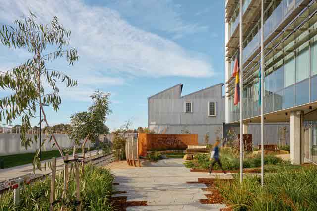 Modern building with glass facade surrounded by landscaped garden and walkways, under a clear blue sky