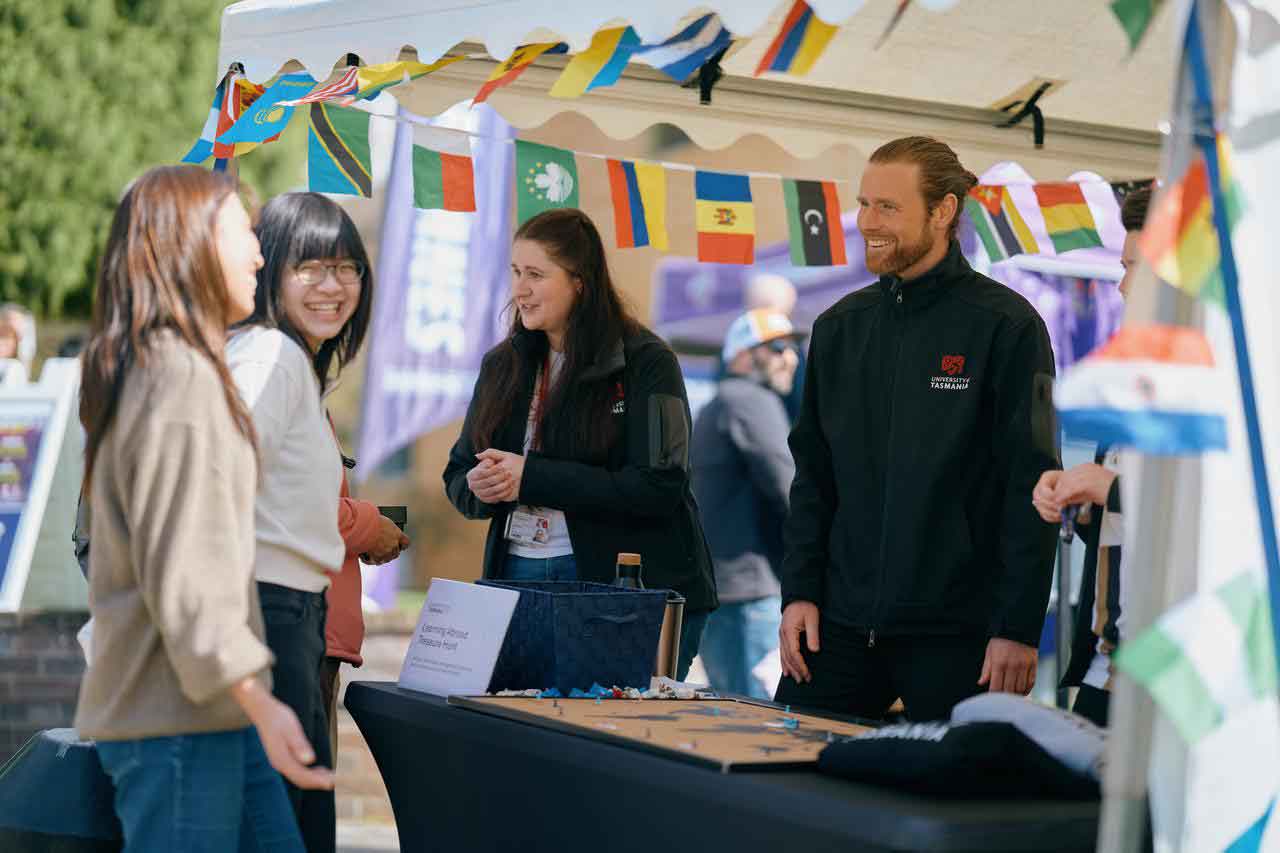 A group of individuals at a booth decorated with international flags, University of Tasmania open day event.