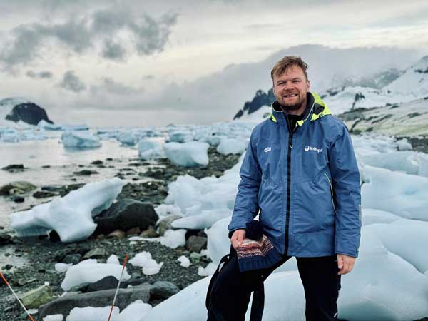 Oscar Vorobjovas-Pinta standing on a rocky shore with icebergs in the water, holding a book, with mountains in the background.